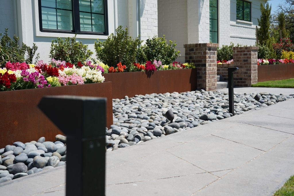 A modern house with white brick walls, large windows, a path bordered by smooth gray stones, colorful flower beds, and rectangular metal planters—this design exemplifies commercial landscape solutions built to withstand the Texas heat.