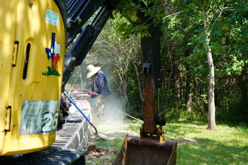 A person in a wide-brim hat uses a power washer near a yellow excavator on grassy ground, surrounded by trees and sunlight.