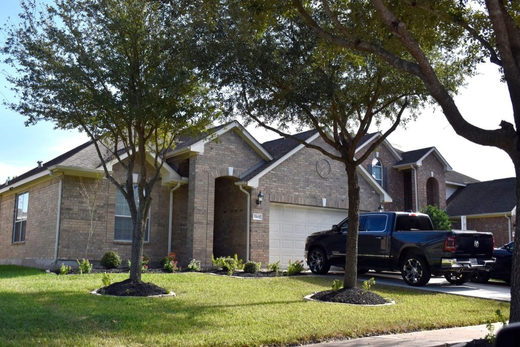 A brick house with a two-car garage and manicured lawn is shaded by trees. A black pickup truck is parked in the driveway. Bright, sunny day.