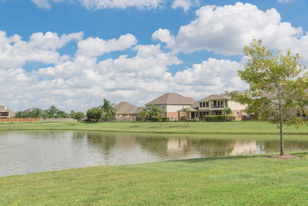 A small lake with green grass surrounds it, a single tree on the right, and several suburban houses in the background under a blue sky with scattered clouds.