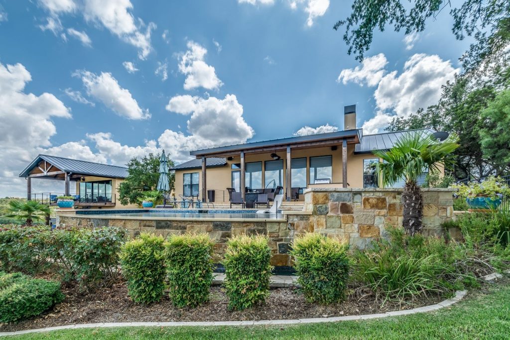 Modern house with stone and stucco walls, large windows, and a covered patio, surrounded by lush landscaping and green shrubs under a bright blue sky with scattered clouds.