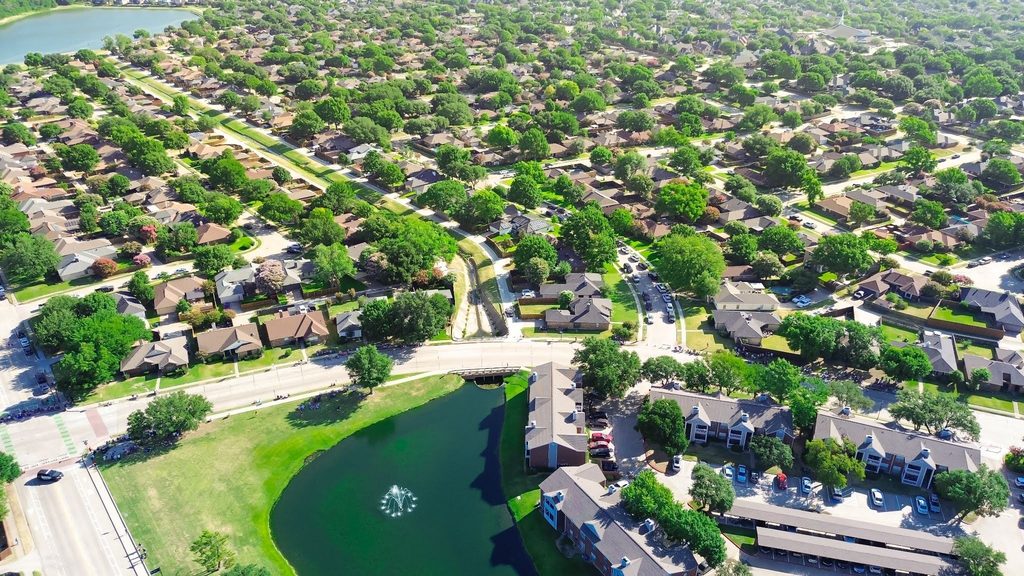 Aerial view of a suburban neighborhood in the fall, with vibrant foliage accenting the green lawns and tree-lined winding streets. The scene showcases well-maintained homes and a small lake with a fountain near an apartment complex, reflecting strict HOA landscape standards that contribute to the neighborhood’s immaculate appearance.