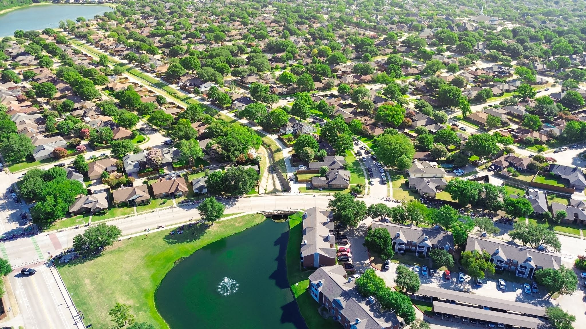 Aerial view of a suburban neighborhood in the fall, with vibrant foliage accenting the green lawns and tree-lined winding streets. The scene showcases well-maintained homes and a small lake with a fountain near an apartment complex, reflecting strict HOA landscape standards that contribute to the neighborhood’s immaculate appearance.
