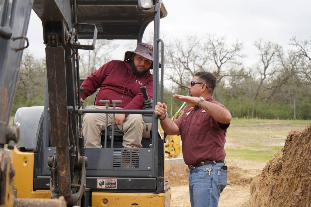 A man in a maroon shirt and hat operates a construction vehicle while another man, also in maroon, gestures and gives directions beside him at a Central Texas worksite with trees in the background, assisting with landscape renovations this fall.