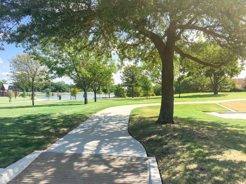 A sunlit concrete path curves through a green park with scattered trees, grassy lawns, and a small pond, all meticulously maintained to meet HOA landscape standards, under a clear blue sky.