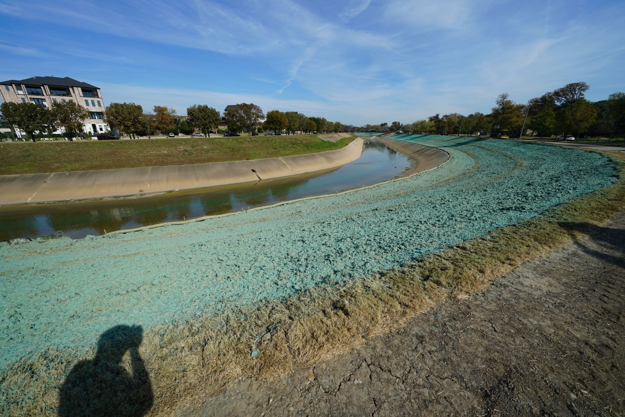 A winding creek lined with concrete walls, with the surrounding dirt banks recently covered in hydroseeding.