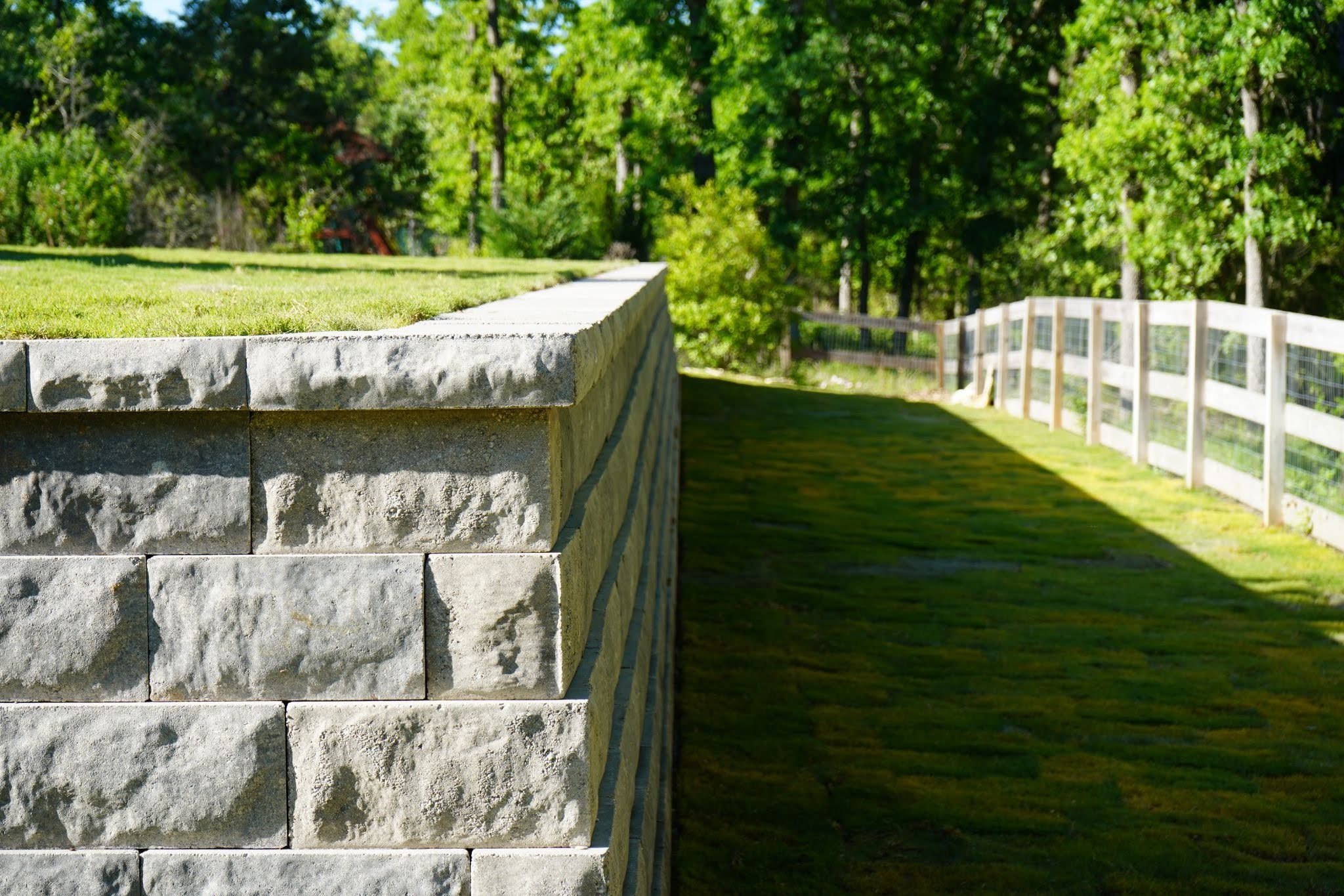 A durable hardscape concrete block retaining wall borders a grass lawn with a wooden fence in the background, showcasing professional Central Texas landscaping.